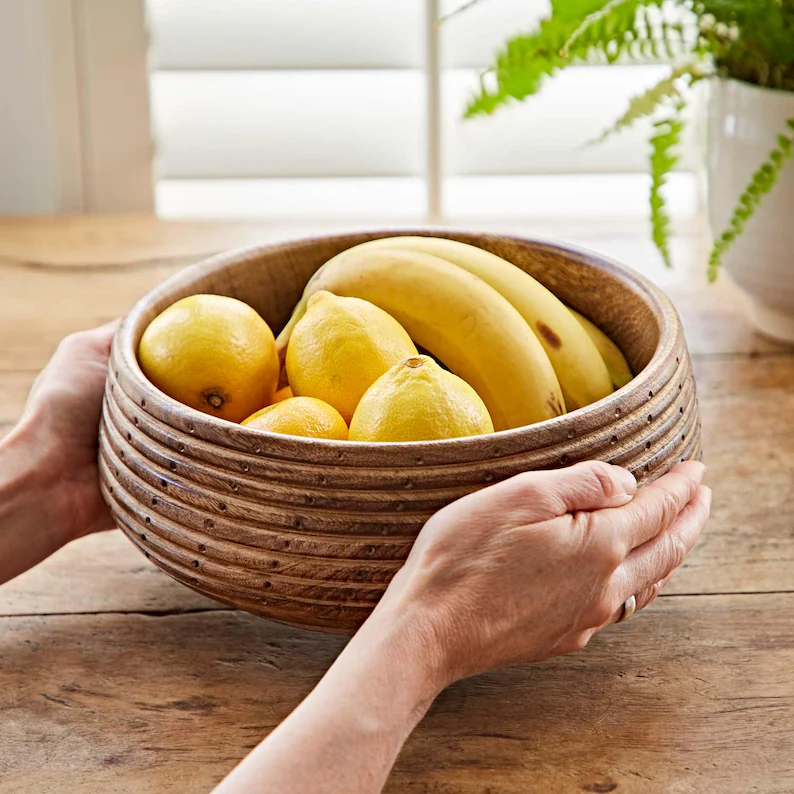 Bol din lemn de mango ținut în mâini, plin cu lămâi și banane, pe o masă de lemn
